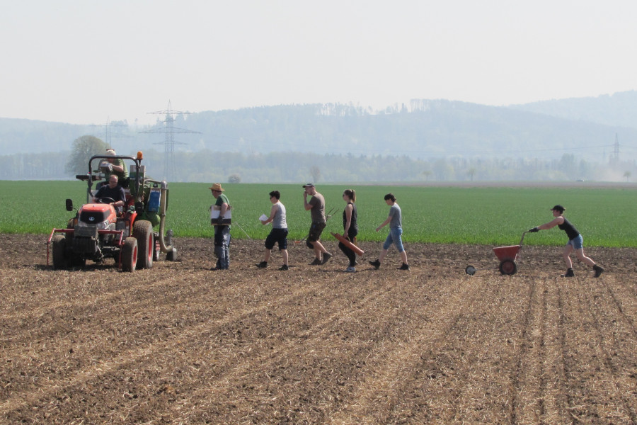 6 Personen gehen in einer Reihe über ein Feldd auf einen kleinen Trecker zu und tragen verschiedene Gegenstände